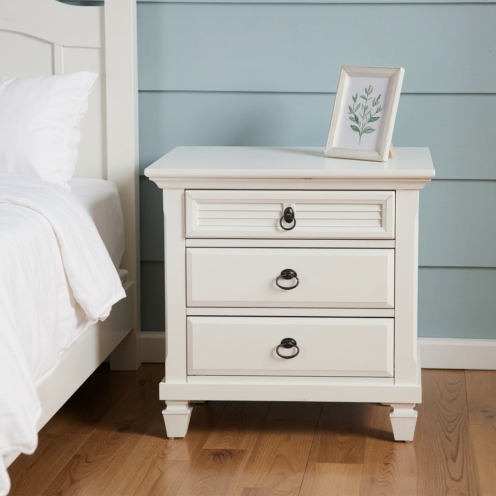 White wood nightstand with three drawers and black ring pull handles next to bed
