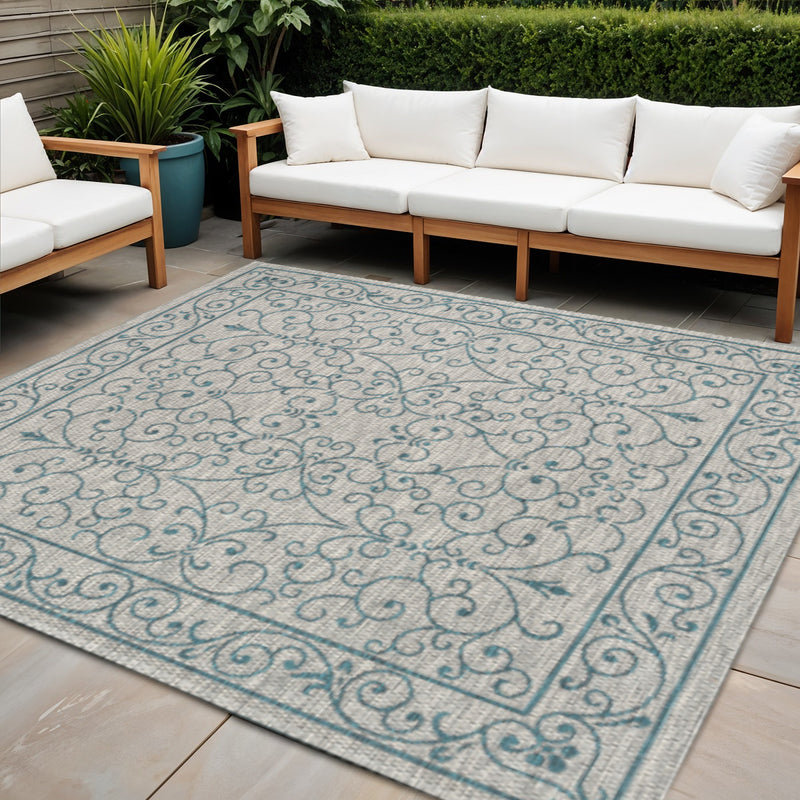Indoor outdoor area rug with large teal floral scroll pattern on light gray background.
