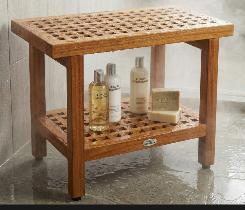 Brown teak shower bench with slatted top, lower shelf, and bath items on shelf