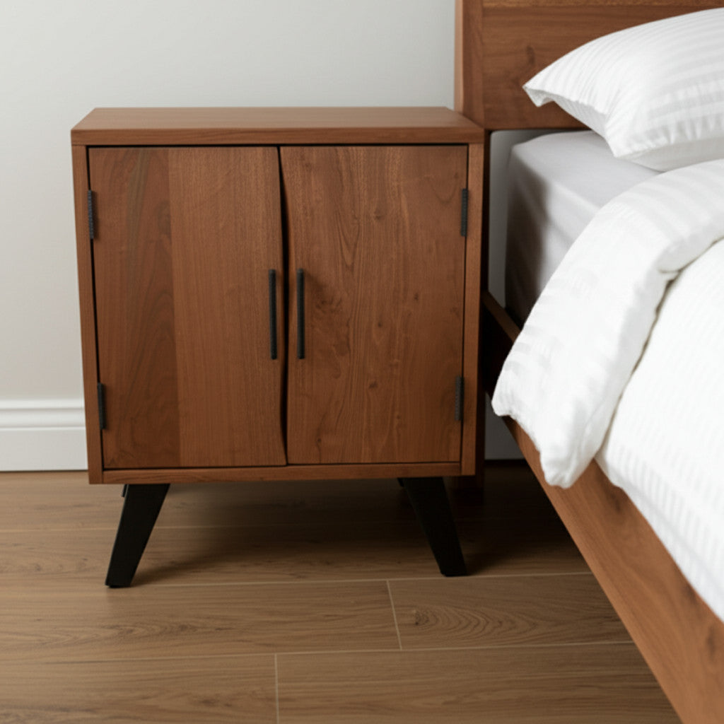 Brown wood nightstand with two doors and dark legs next to a bed