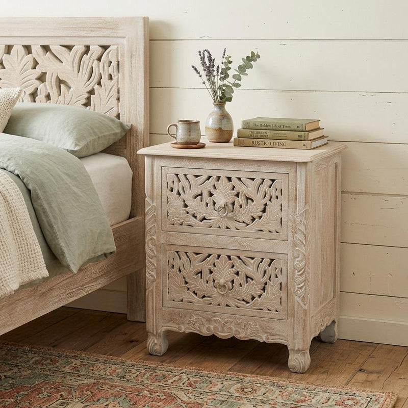 Distressed white carved floral nightstand with two drawers, shown beside a bed