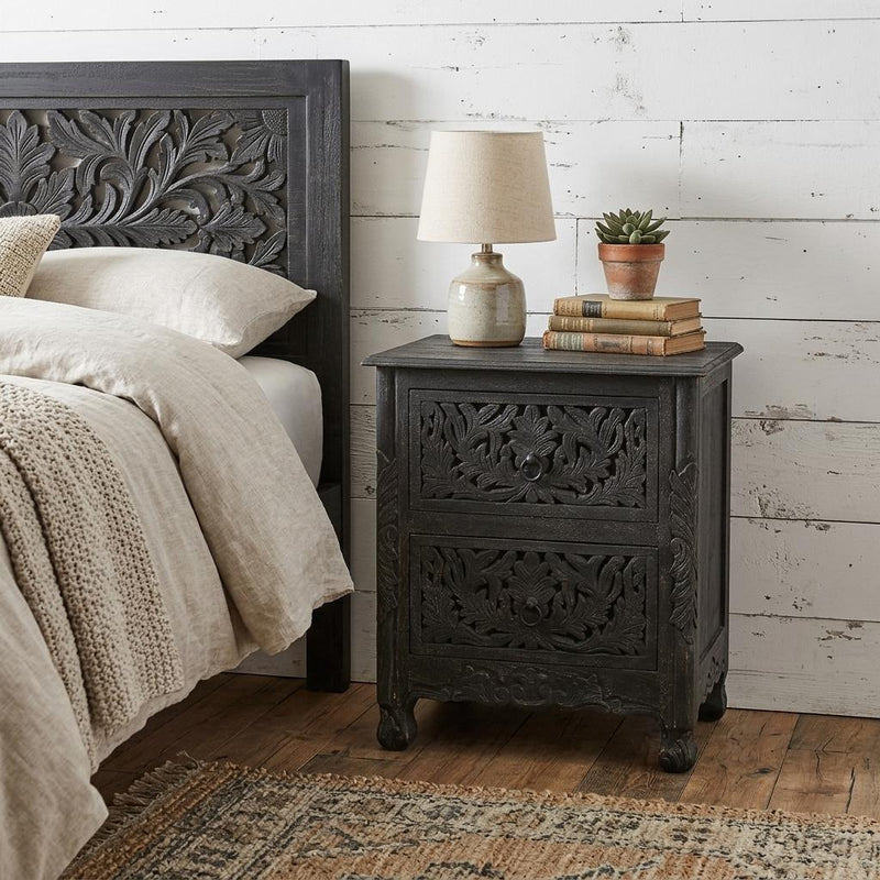 Distressed black wood nightstand with carved drawer fronts beside a bed