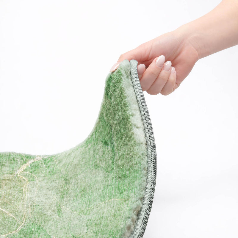 Hand lifting green abstract shag area rug corner to show thick pile and gray binding.