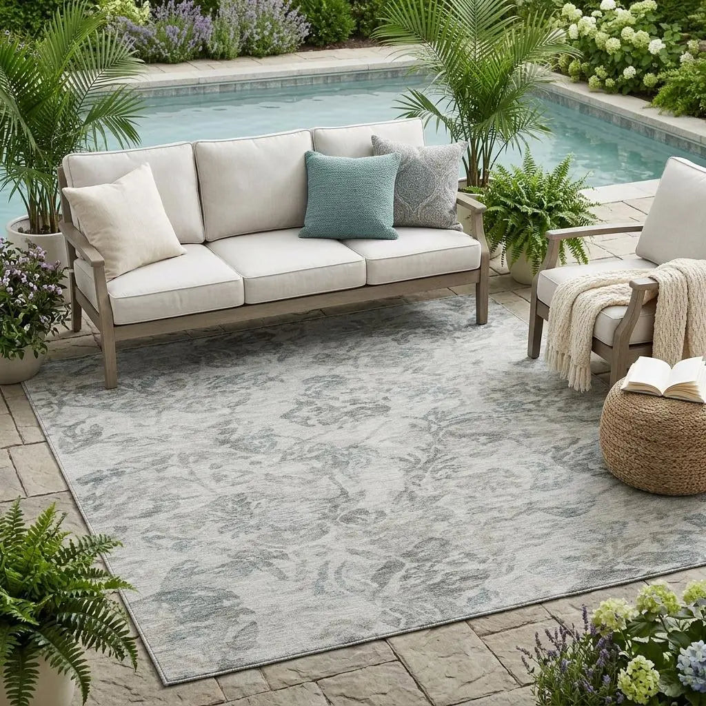 Outdoor area rug in soft gray and blue floral pattern, complemented by a beige sofa and greenery by a pool.