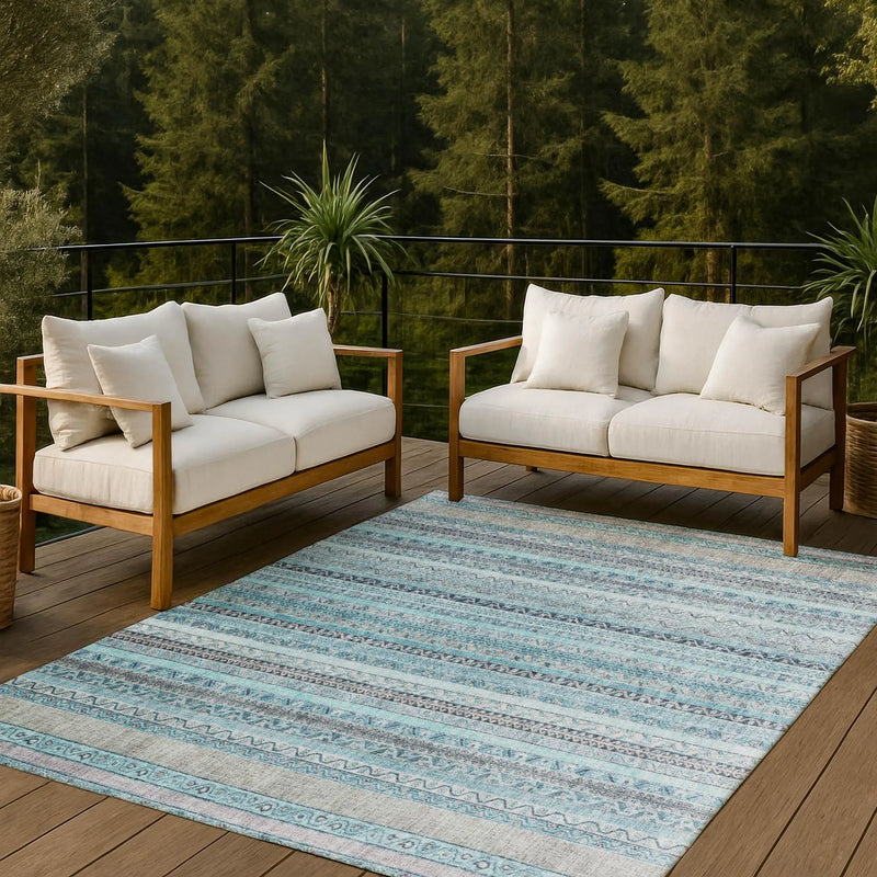 Light blue boho area rug on a wooden patio with beige cushioned sofas and greenery in the background.