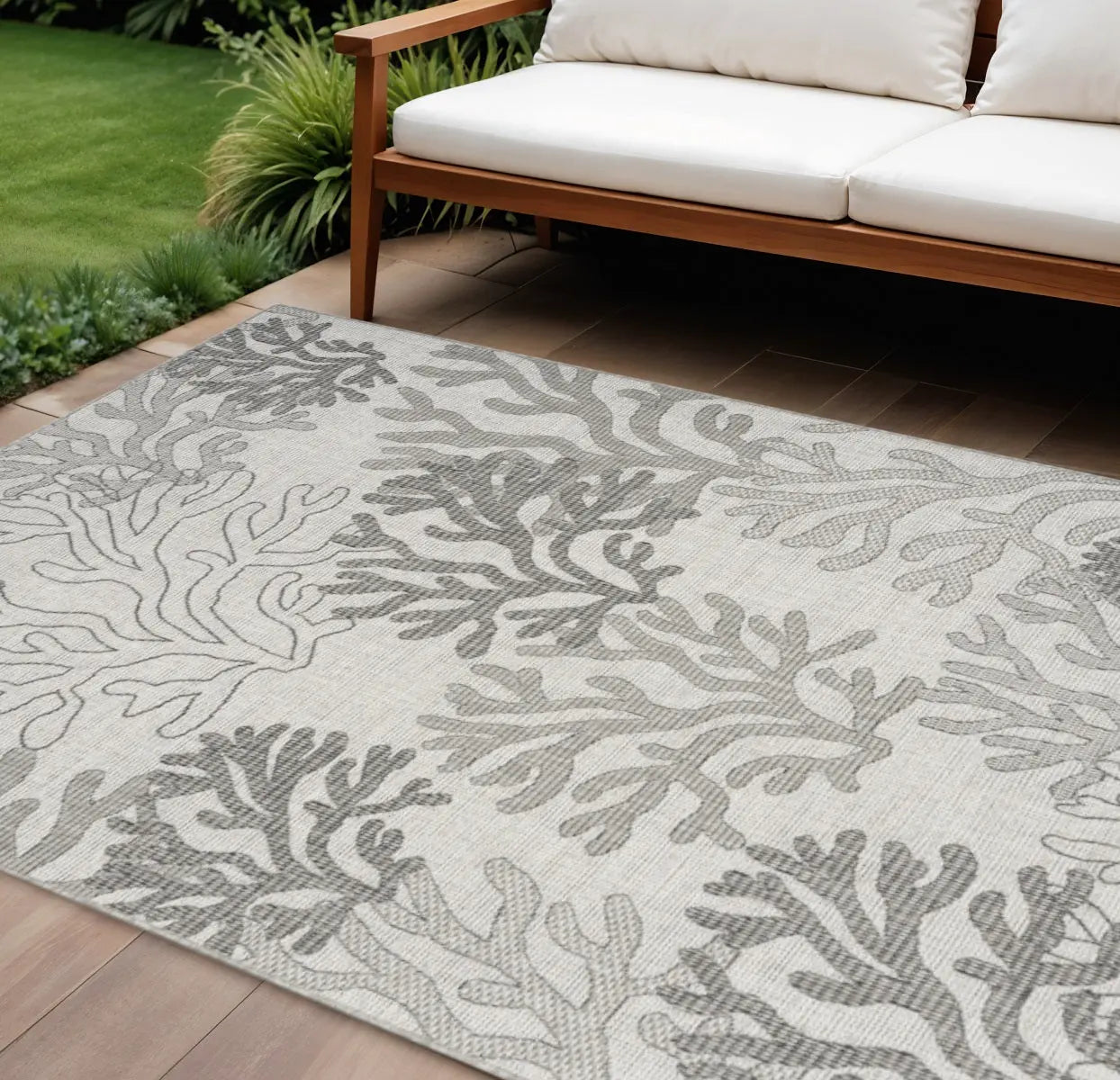 Light gray coral-patterned outdoor rug on a wooden patio beside a plush white sofa with green landscaping.