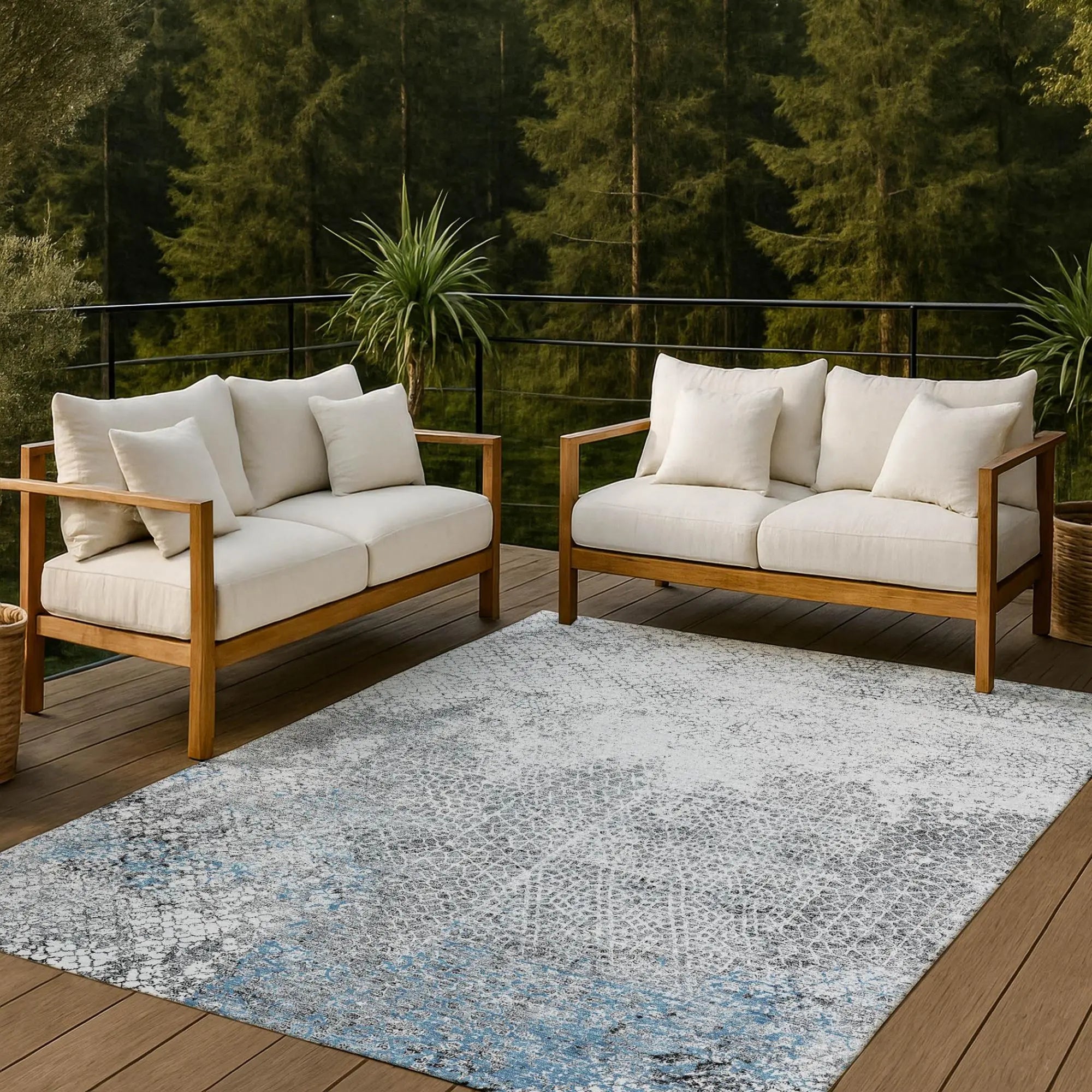 Light gray and blue abstract area rug on a wooden patio with beige outdoor sofas and green foliage background.