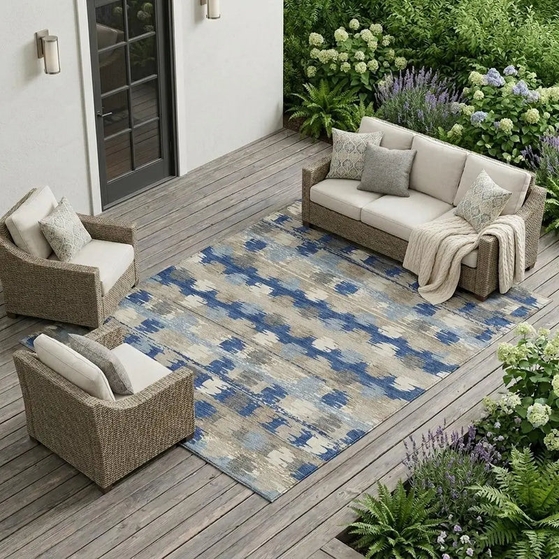 Blue and beige patterned outdoor rug on a wooden patio with wicker furniture and greenery.