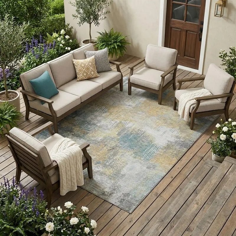 Beige outdoor sofa set with wooden accents, blue and yellow patterned rug, surrounded by greenery and flowers.