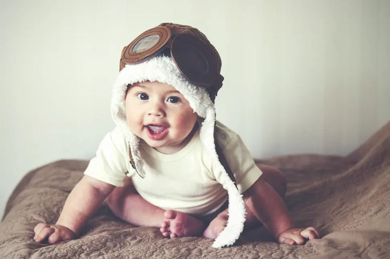 Adorable baby wearing a brown aviator hat with goggles, white fluffy trim, on a textured beige blanket.