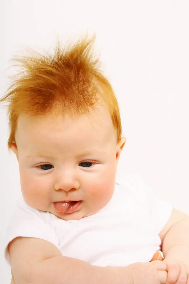 Cute baby with spiky red hair, wearing a white short-sleeve shirt, showcasing adorable expression and tongue out.