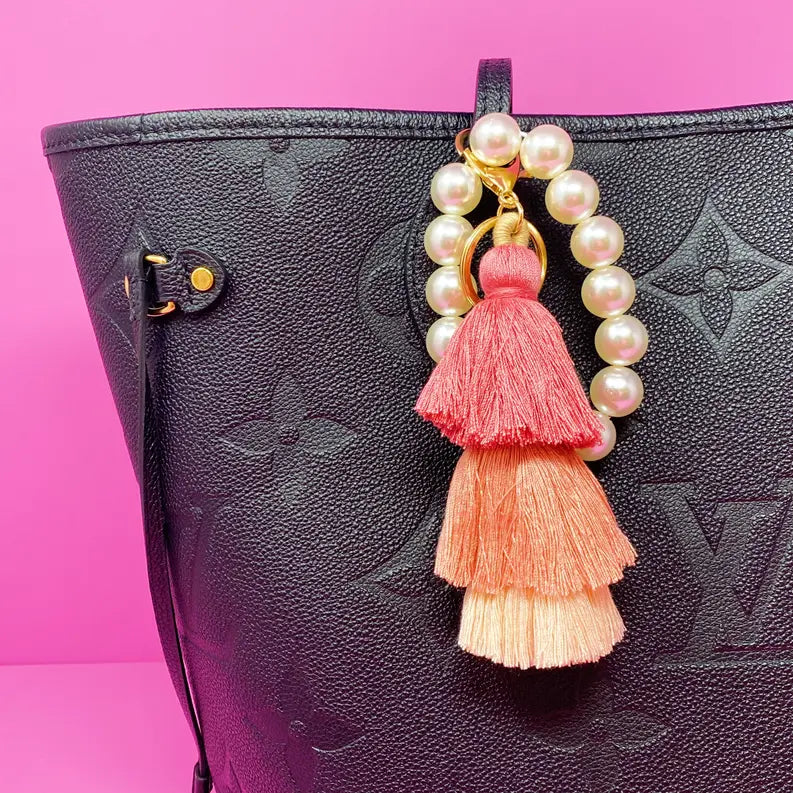 Black textured leather tote bag with a pink ombre tassel charm and pearl accents on a pink background.