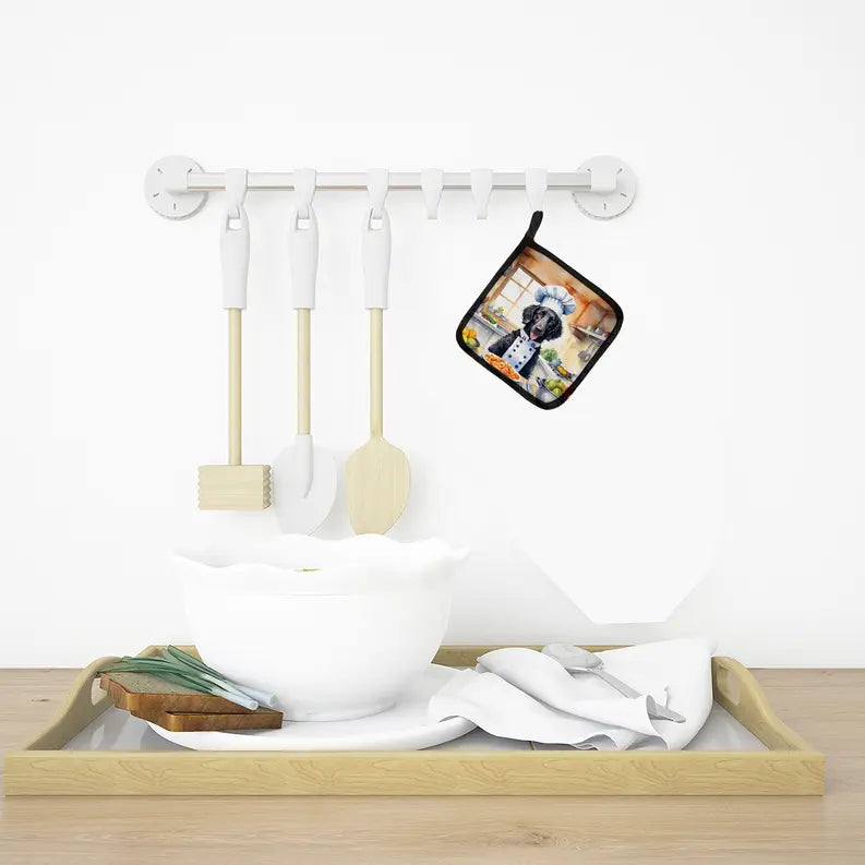 Black and white cotton pot holder featuring a cute dog design, hanging alongside white kitchen utensils on a wooden tray.
