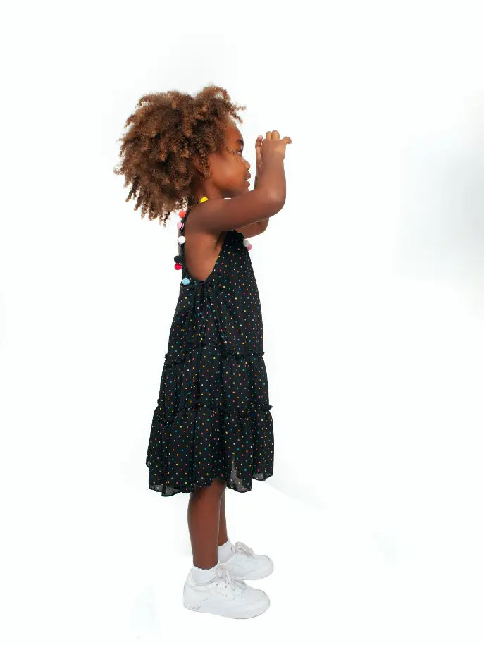 Black polka dot tiered dress for kids, styled with colorful pom-poms, perfect for summer outings.