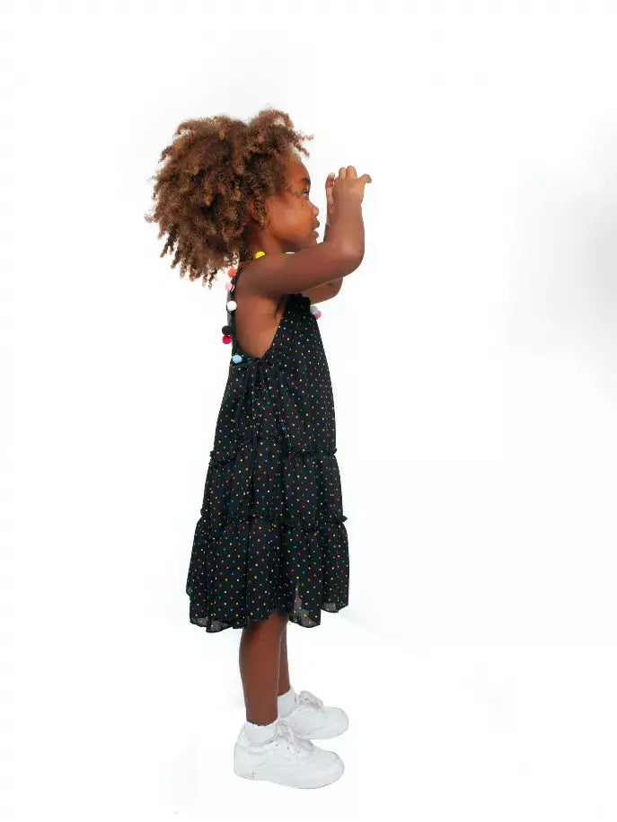 Black tiered dress with colorful polka dots for kids, styled with white sneakers and playful accessories.