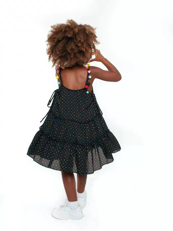 Black polka dot tiered dress with colorful pom-poms on shoulders, perfect for kids' summer fashion.