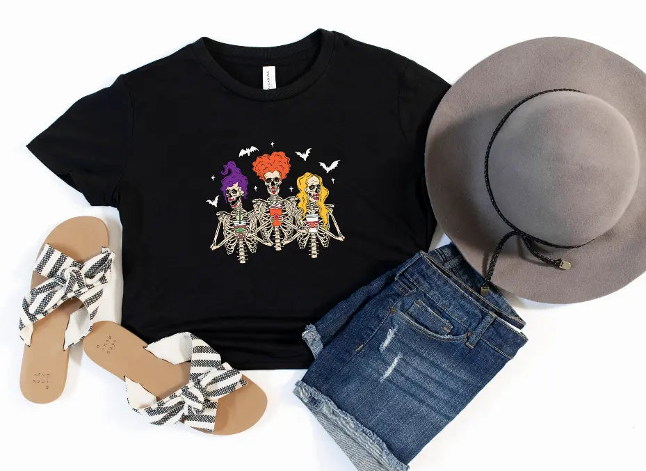 Black t-shirt with colorful skeleton graphic, paired with denim shorts, wide-brim hat, and striped sandals.
