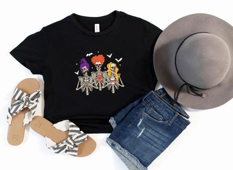 Black t-shirt with colorful skeleton graphic, paired with denim shorts, wide-brim hat, and striped sandals.