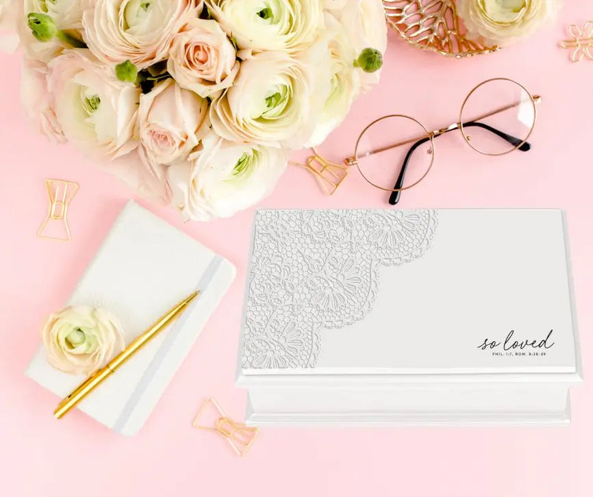 Elegant white lace journal with "so loved" in stylish font, surrounded by pale pink roses and gold accessories.
