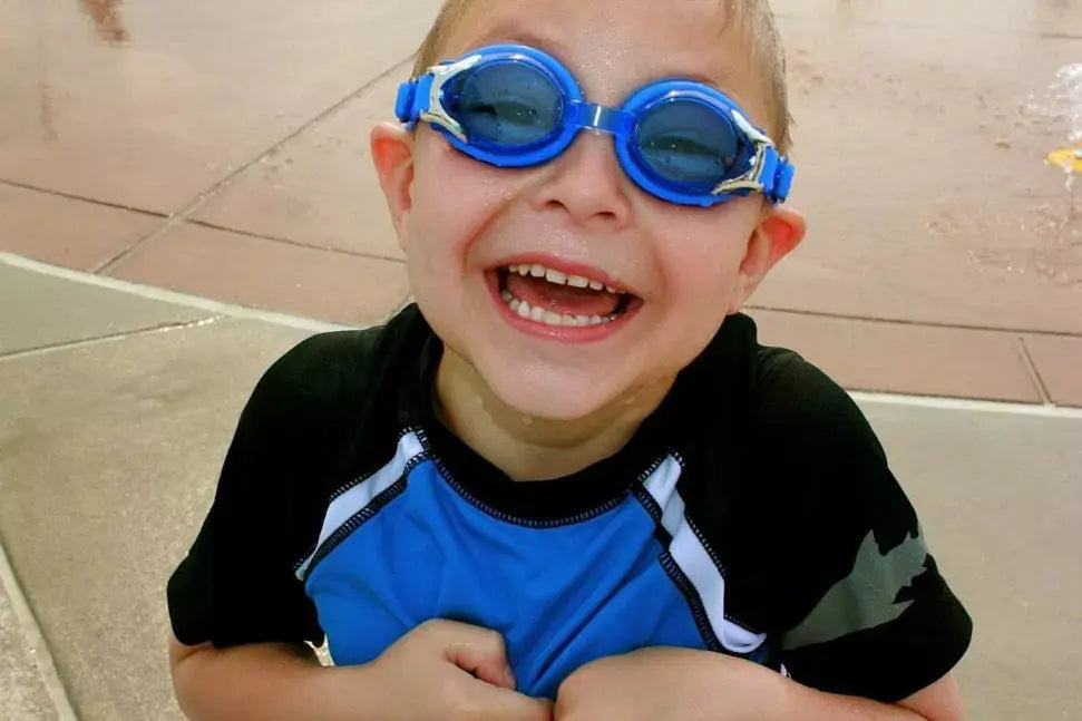 Smiling boy in blue swimming goggles and a black-blue rash guard enjoying water play.