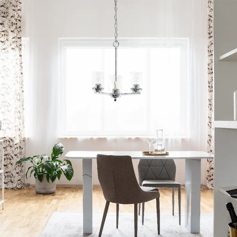 Modern chrome chandelier in a bright dining room with a white table, gray upholstered chair, and green plants.