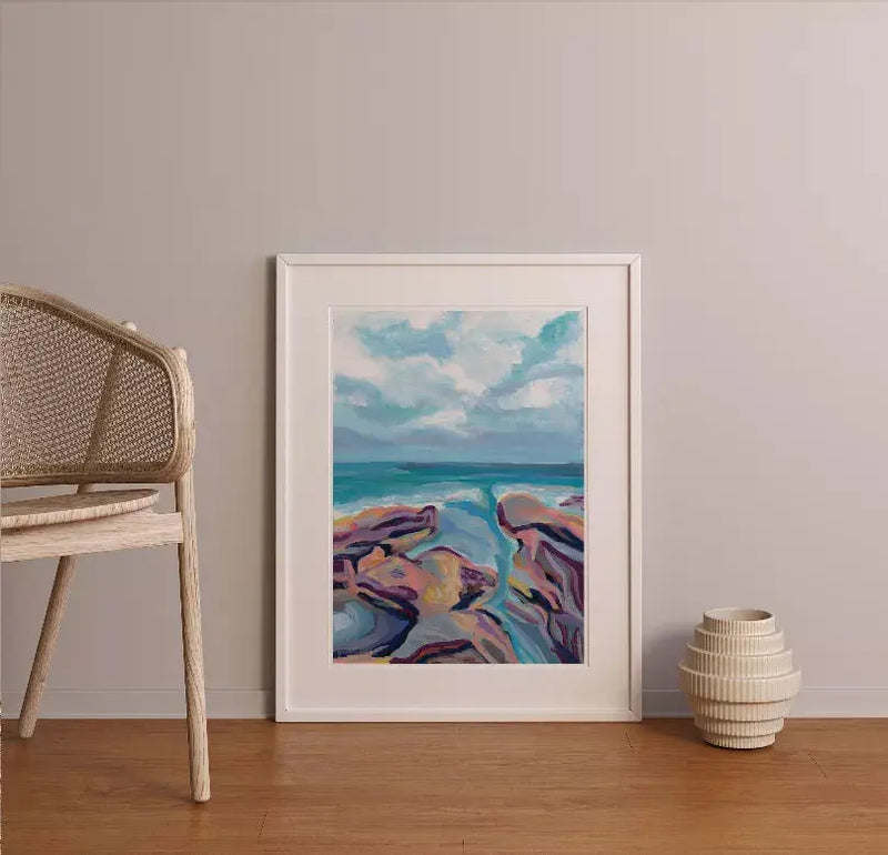 Framed abstract coastline painting in blue, purple, and coral on a wall, complemented by a woven chair and ceramic vase.
