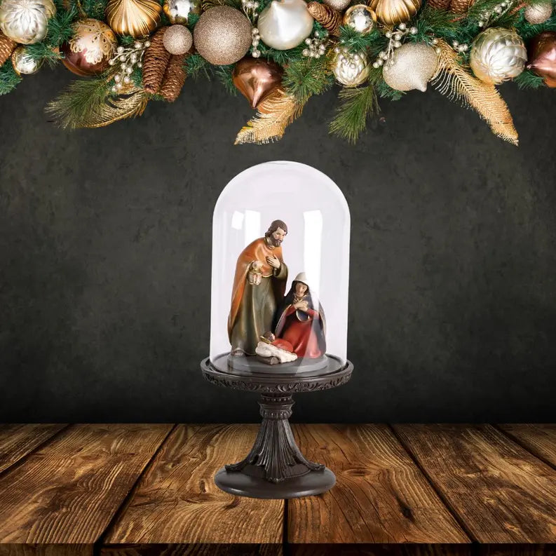 Nativity scene figurine under glass dome on ornate black pedestal with gold and green holiday decor in background.