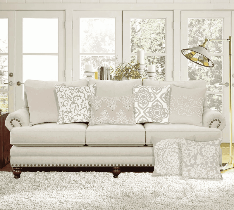 Beige upholstered couch with decorative pillows in white and taupe, featuring elegant patterns in a bright, airy living room.