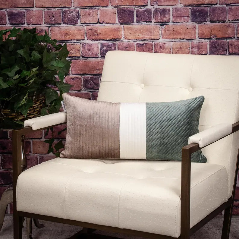 Cream and teal velvet decorative pillow with quilted design, resting on a modern lounge chair against a brick wall.