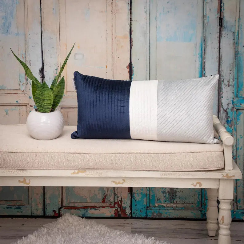 Navy blue and silver quilted lumbar pillow on beige bench, with a green plant in a white vase and rustic wooden backdrop.