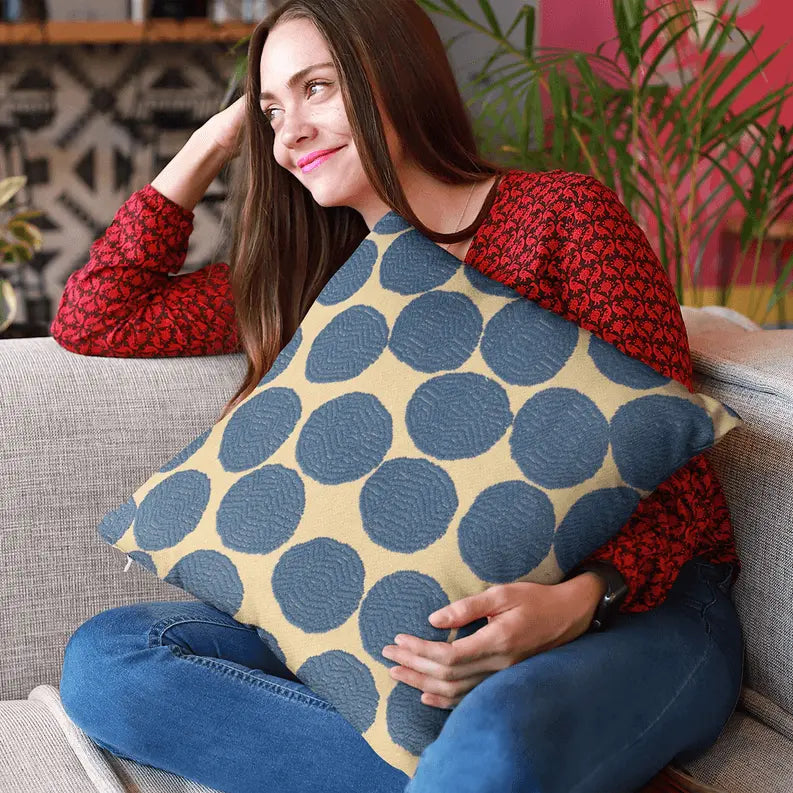 Cream decorative pillow with navy blue textured dots, showcased by a woman in a red patterned blouse on a gray couch.