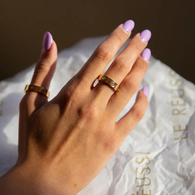 Gold minimalist rings with elegant stone accents worn on a hand with lavender nails, set against delicate tissue paper.