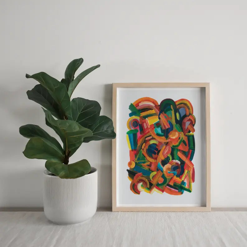 Colorful abstract art print in a natural wood frame beside a lush green fiddle leaf fig in a white textured pot.