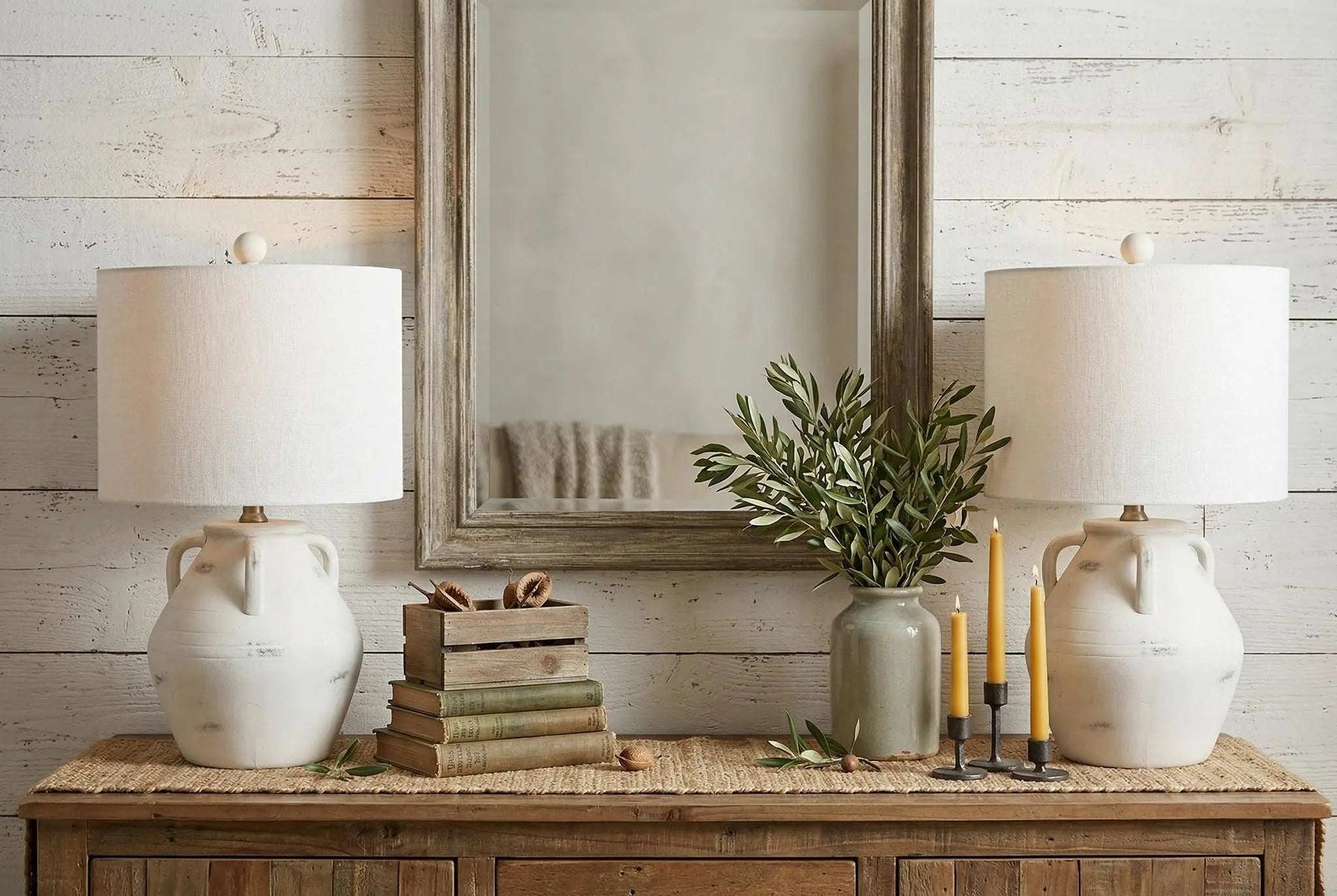 White ceramic table lamps with linen shades, styled with rustic decor, olive branches, and candles on a wooden console.