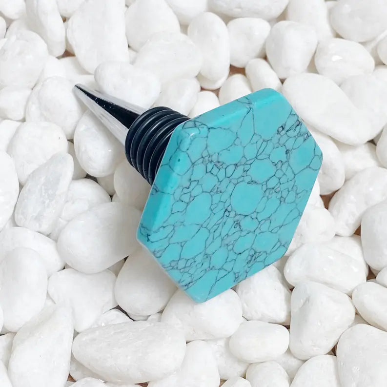 Turquoise hexagon wine stopper with black rubber base, elegantly designed on a white pebble background.