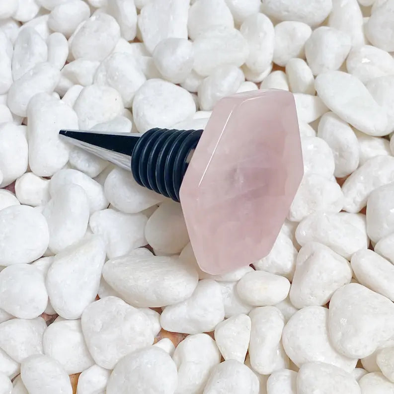 Rose quartz wine stopper with a sleek black metal base, resting on smooth white pebbles. Elegant and stylish addition for wine lovers.