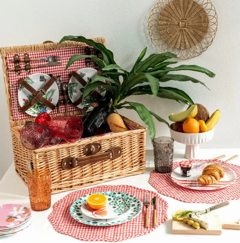 Wicker picnic basket with red checkered lining, plates, glasses, and fresh fruits, set decoratively on a white table.