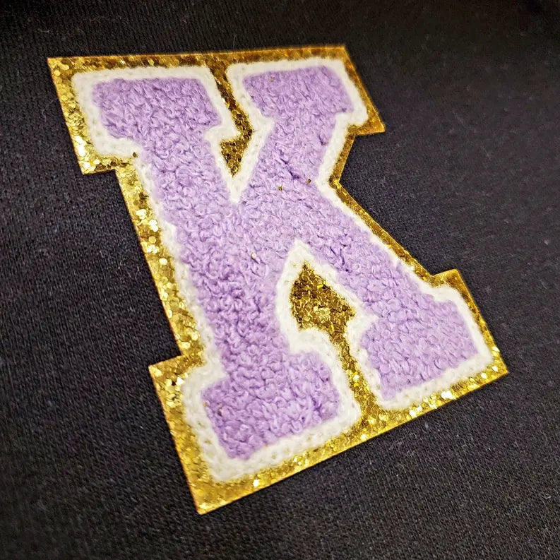 Purple "K" patch with textured fabric and gold glitter trim on a black background. Ideal for custom apparel or accessories.